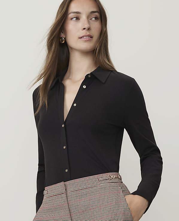 A woman in a black collared shirt and patterned pants stands against a plain background.