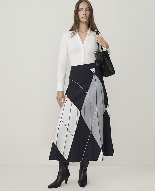 A woman in a white shirt, patterned skirt, and black boots stands against a plain white background, holding a black handbag.