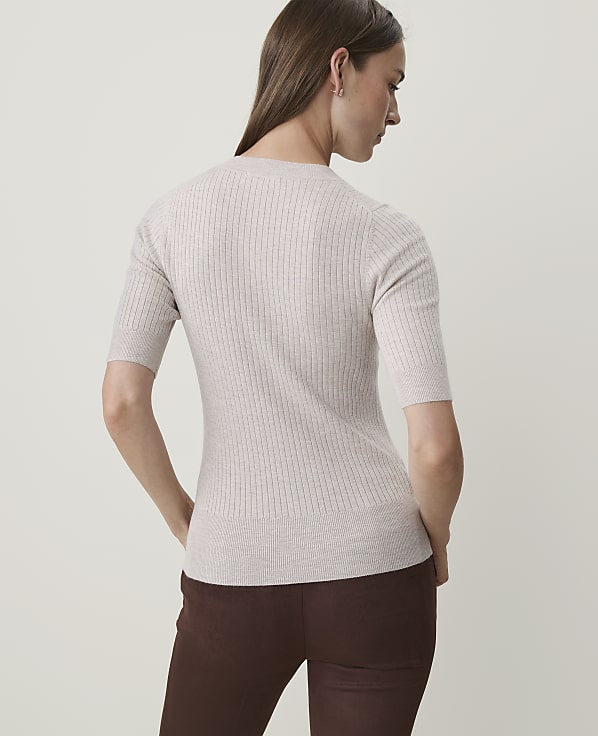 A woman in a light-colored ribbed sweater and brown pants stands against a plain, light background.