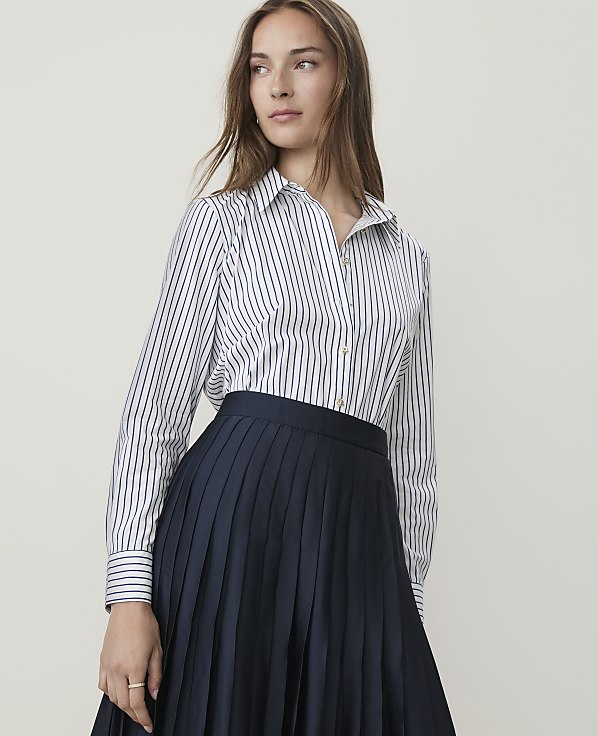 A woman in a striped shirt and pleated skirt stands against a plain background.
