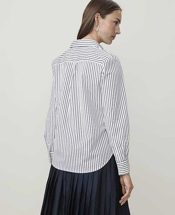 A woman in a striped shirt and pleated skirt stands against a plain background.