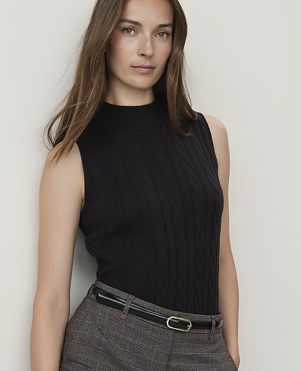 A woman in a black cable-knit sleeveless turtleneck and plaid pants stands against a plain white background.