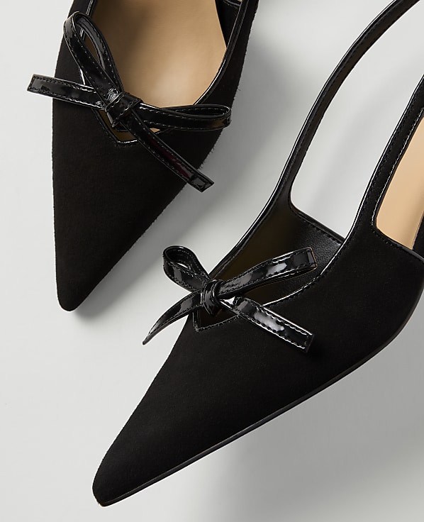 Two black suede slingback heels with patent leather bows are arranged on a white background.