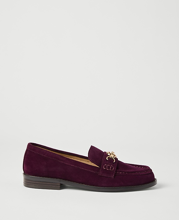 A deep purple suede loafer with a gold chain detail rests on a plain white background.