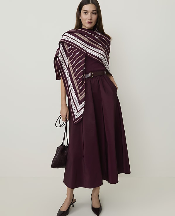 A woman in a burgundy dress and patterned scarf stands against a plain, light background, holding a dark handbag.