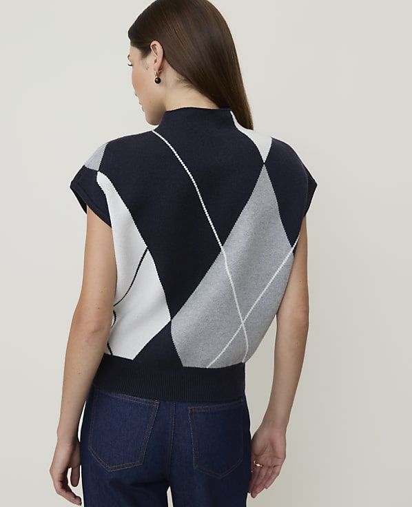 A woman in a navy, white, and gray argyle sweater vest and dark jeans stands against a plain, light gray background.