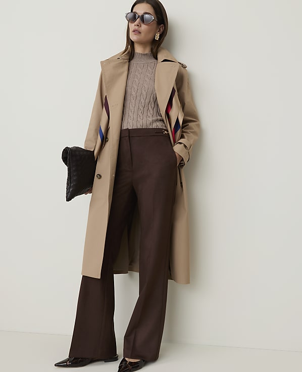 A woman in a trench coat and wide-leg pants stands against a plain white background, holding a clutch.