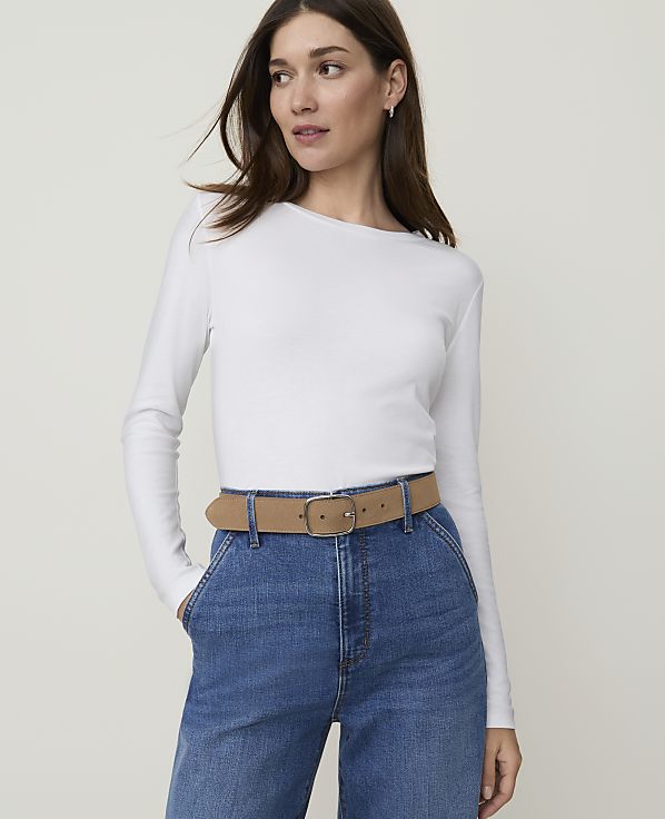 A woman in a white long-sleeved shirt and blue jeans with a tan belt stands against a plain white background.