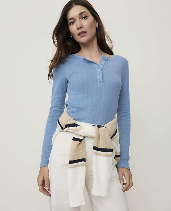 A woman in a blue henley shirt and white pants stands against a plain, light gray background, with a striped sweater tied around her waist.