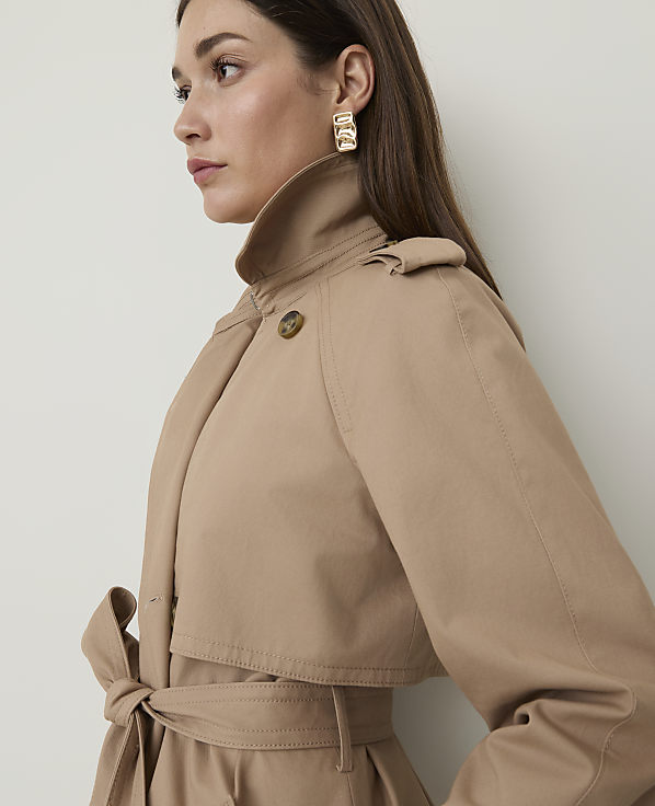 A woman in a tan trench coat with a tie at the waist is shown in profile against a plain white background.