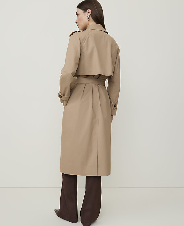 A woman in a tan trench coat and brown pants stands against a plain, light background.