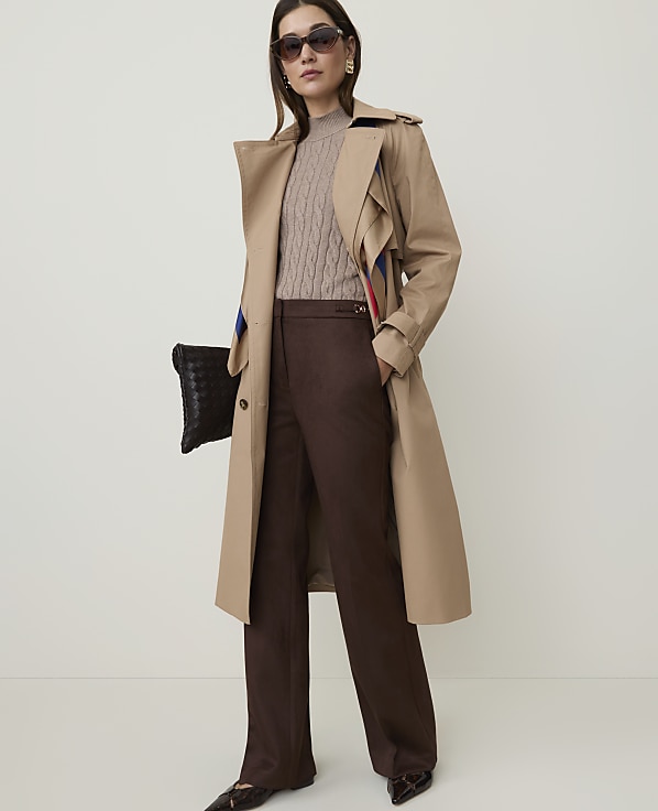 A woman in a tan trench coat and brown pants stands against a plain, light-colored background, holding a black clutch.