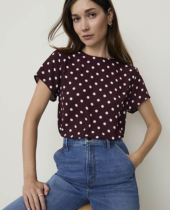 A woman in a burgundy polka dot top and blue jeans poses against a plain white background.