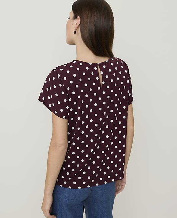 A woman in a burgundy polka dot top and blue jeans stands against a plain white background.