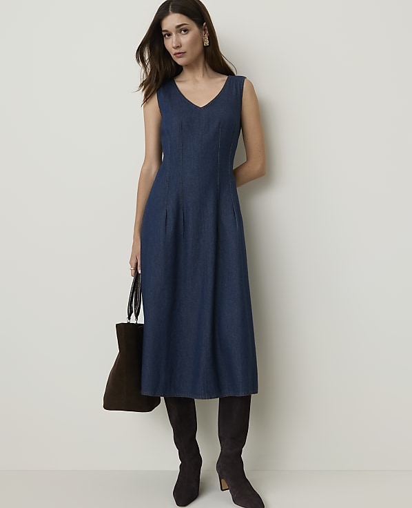A woman in a denim dress and boots stands against a plain background, holding a brown bag.