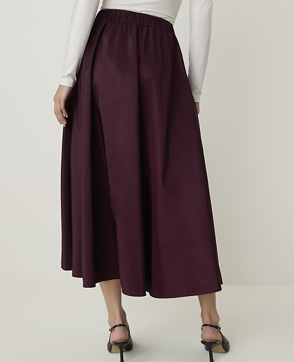 A person wearing a burgundy midi skirt and a white long-sleeved shirt stands against a plain light background.