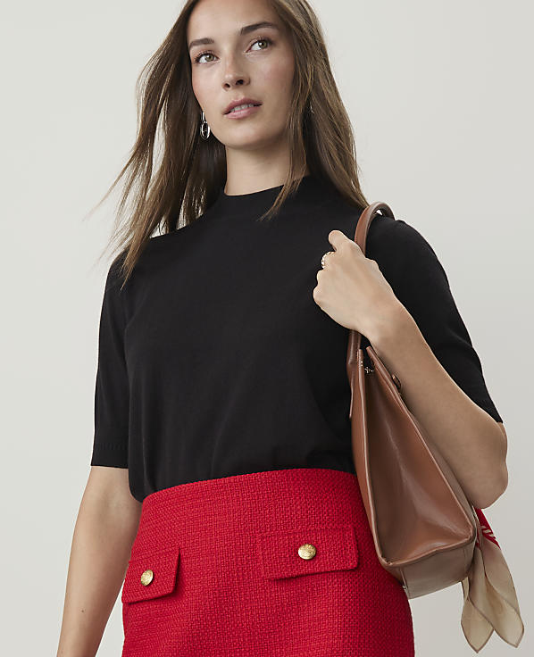 A woman in a black top and red skirt carries a brown purse against a plain background.