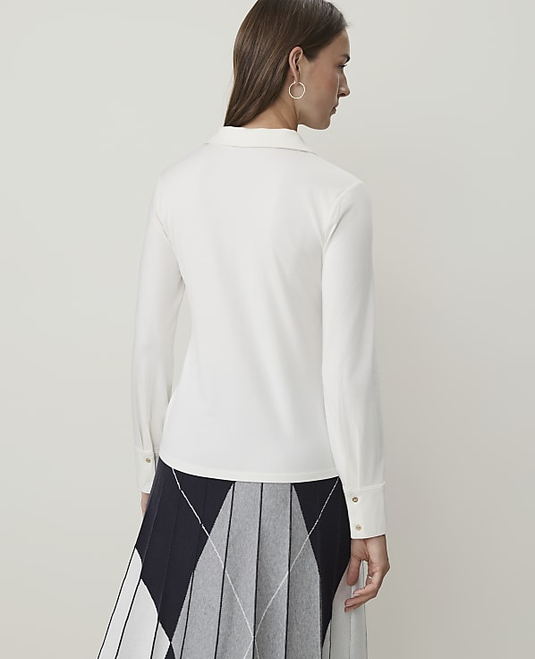A woman in a white collared shirt and a pleated skirt stands against a plain white background.