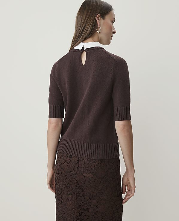 A woman in a brown sweater and lace skirt stands against a plain white background.