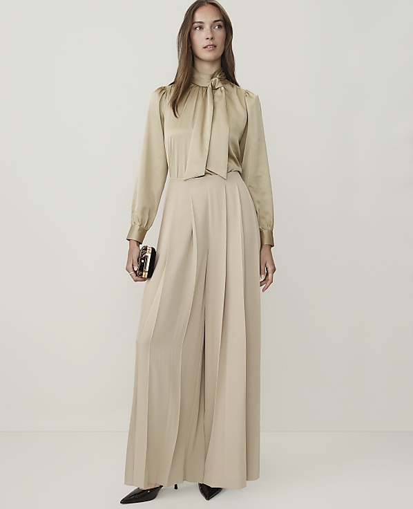 A woman in a champagne-colored satin blouse and flowing wide-leg pants stands against a plain, light background, holding a small, ornate clutch.