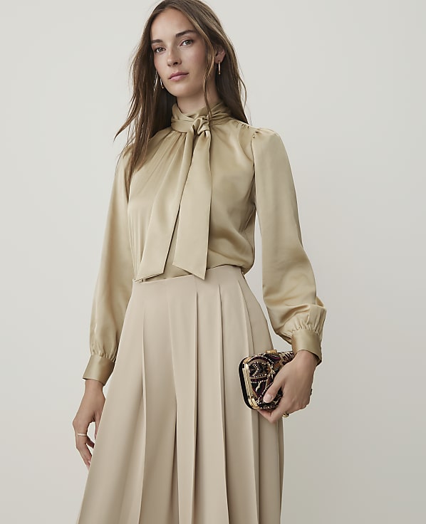 A woman in a champagne-colored silk blouse and wide-leg pants holds a jeweled clutch against a plain background.