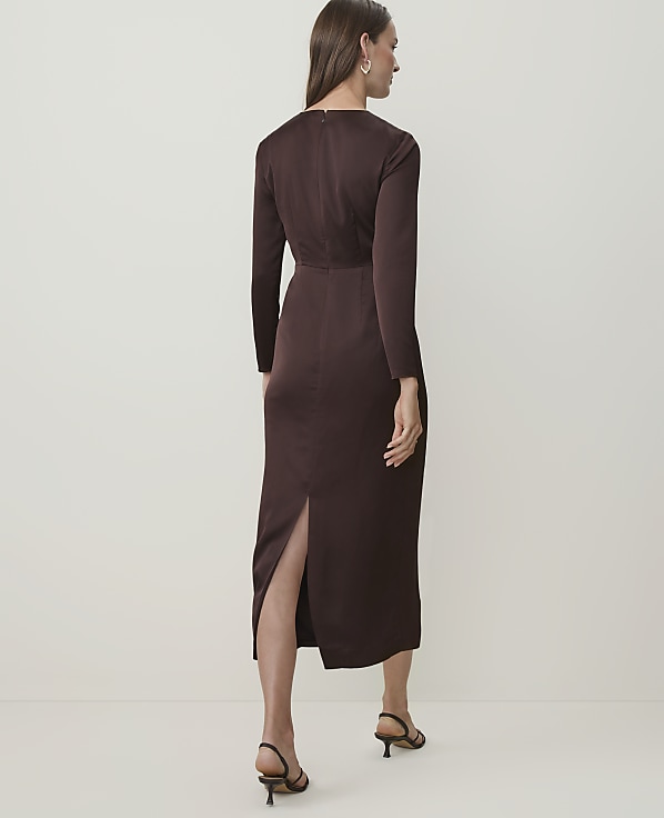 A woman in a brown dress stands against a plain, light-colored background.