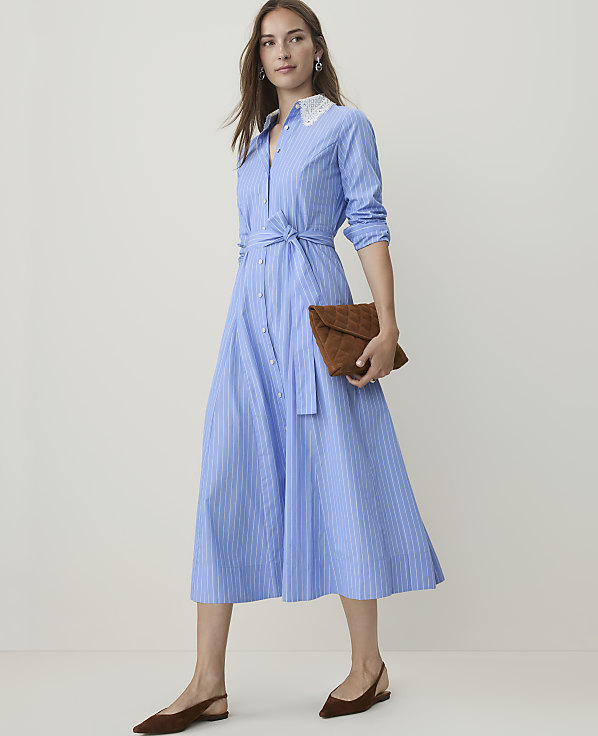 A woman in a blue and white striped dress with a white lace collar and brown flats stands against a plain, light background, holding a brown quilted clutch.