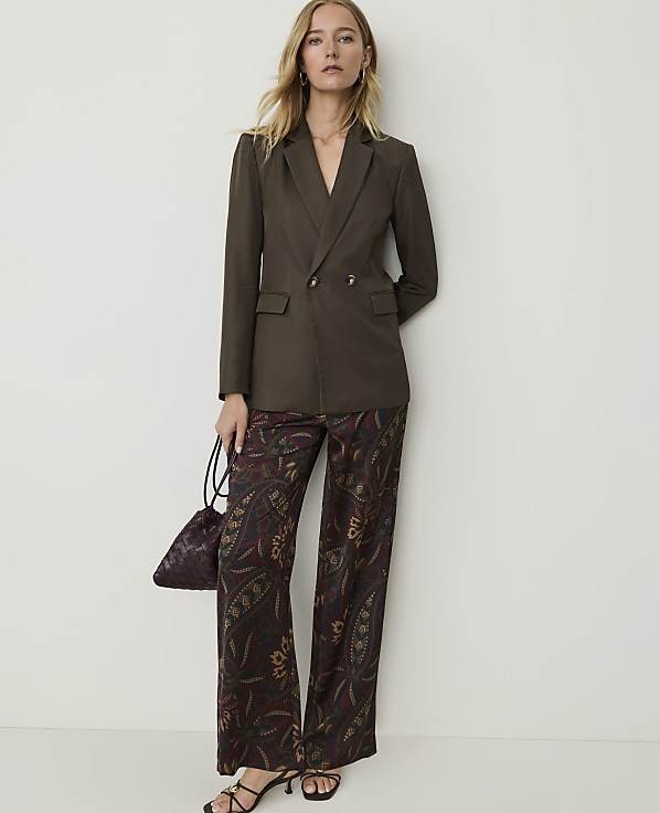 A woman in a brown blazer and patterned pants stands against a plain white wall, holding a small patterned bag.