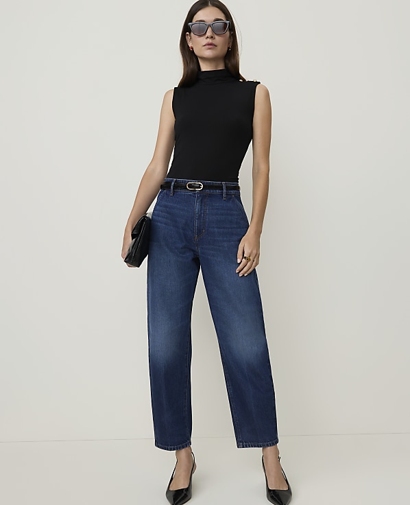 A woman in a black sleeveless top and high-waisted jeans stands against a plain white background, holding a black clutch.