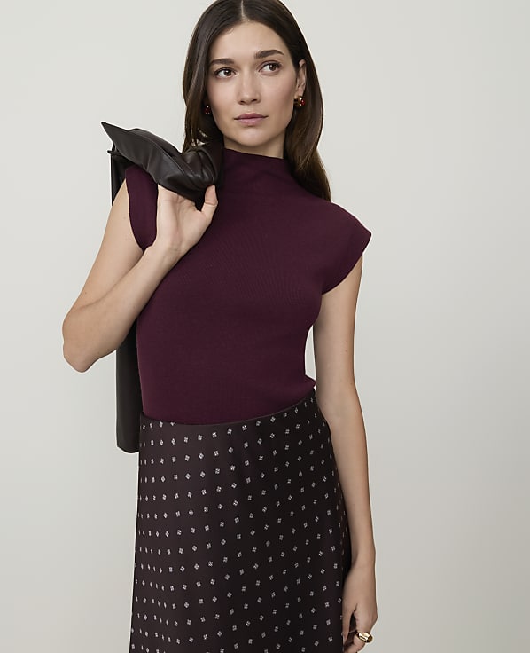 A woman in a burgundy mock-neck top and patterned skirt stands against a plain gray background, with a dark jacket draped over her shoulder.