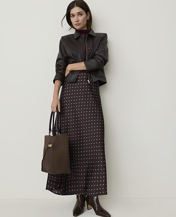 A woman in a patterned skirt and leather jacket stands with a handbag against a plain wall.