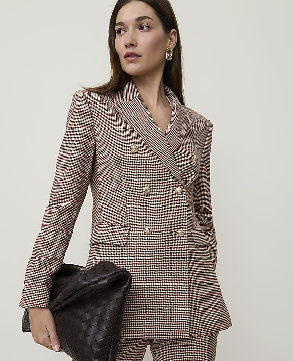 A woman in a patterned blazer and pants holds a woven clutch in front of a plain background.