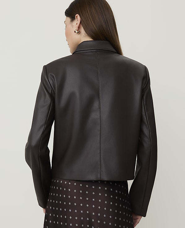 A woman in a dark brown faux leather jacket and patterned skirt stands against a plain, light background.