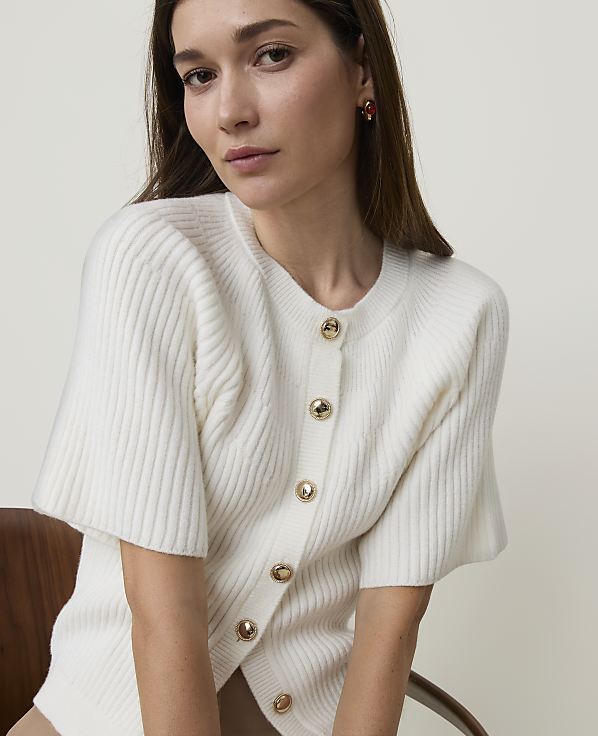 A woman in a white ribbed cardigan with gold buttons sits in front of a blurred wooden chair.