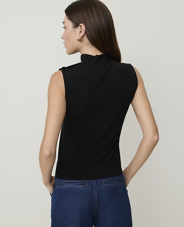 A woman in a black sleeveless turtleneck and blue jeans stands against a plain white background.