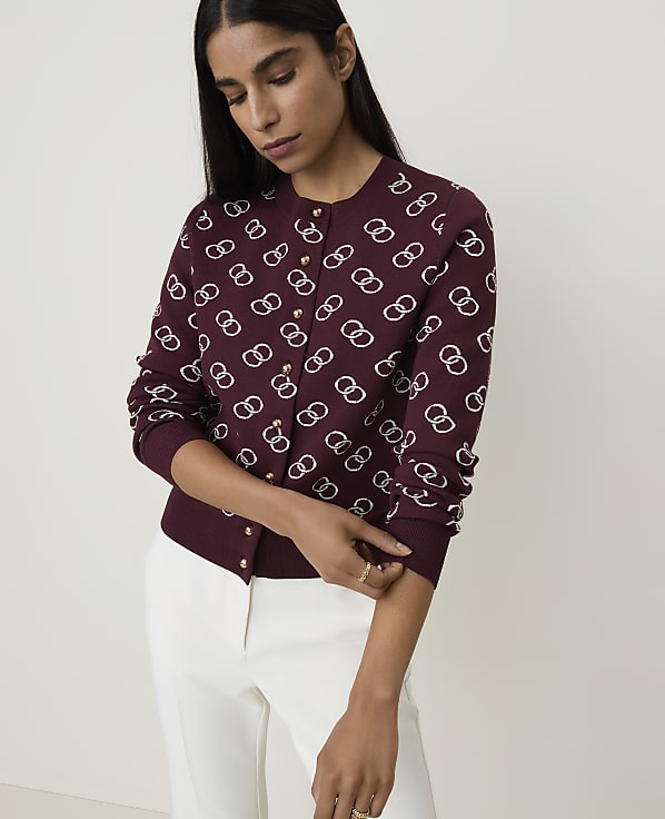 A woman in a patterned burgundy cardigan and white pants stands against a plain white background.