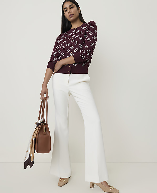 A woman in a patterned burgundy cardigan and white flared pants stands holding a brown handbag against a plain white background.