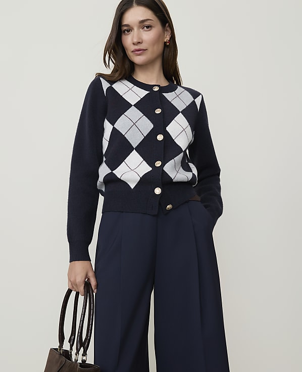 A woman in a navy argyle cardigan and wide-leg navy pants stands against a plain, light background, holding a brown handbag.