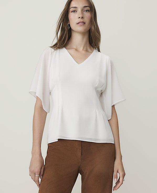 A woman in a white v-neck blouse and brown pants stands against a plain, light gray background.