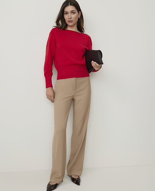 A woman in a red sweater and tan pants holds a black clutch against a plain white background.