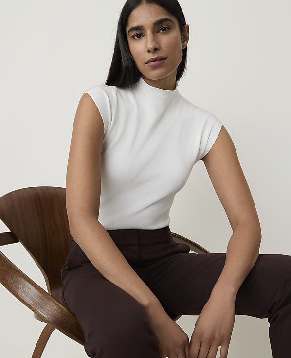 A woman in a white mock-neck top and brown pants sits on a wooden chair against a plain, light background.