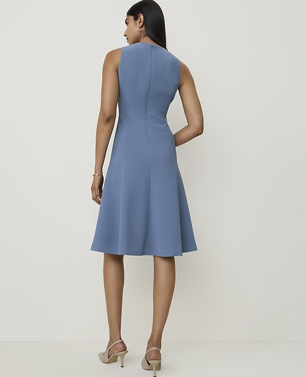 A woman in a blue sleeveless dress stands against a plain, light-colored background.
