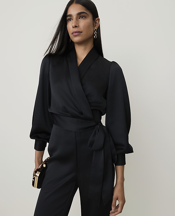 A woman in a black satin jumpsuit with a wrap top and puffed sleeves stands against a plain, light background, holding a small clutch.