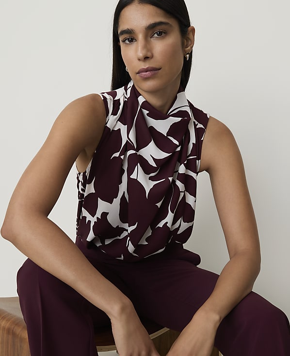 A woman in a patterned blouse and maroon pants sits in front of a plain white background.