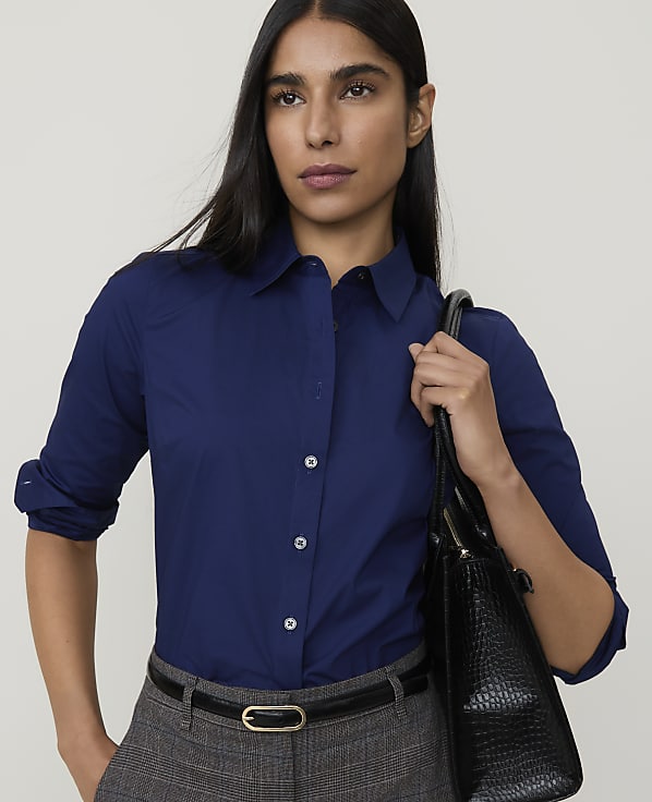A woman in a navy blue collared shirt and grey pants, carrying a black handbag, stands against a plain grey background.
