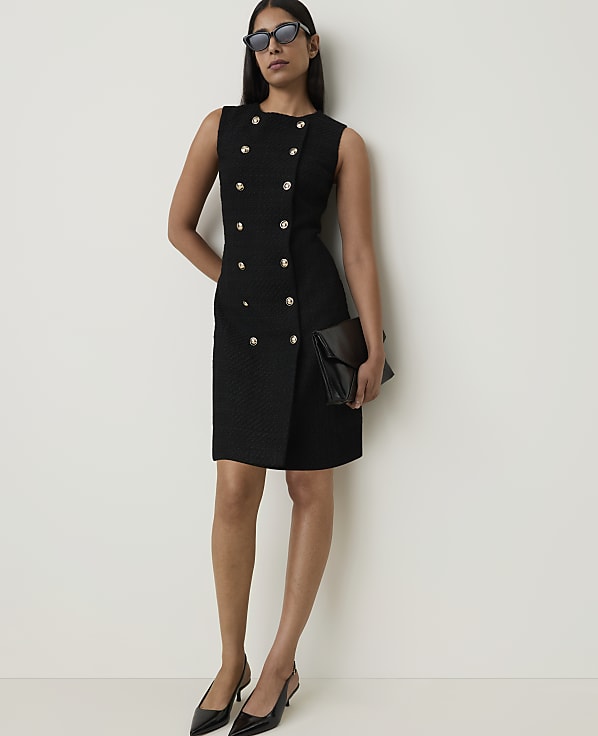 A woman in a black double-breasted dress and sunglasses stands against a plain white background, holding a black clutch.