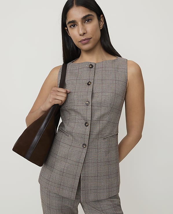 A woman in a plaid vest and pants carries a brown bag against a plain background.