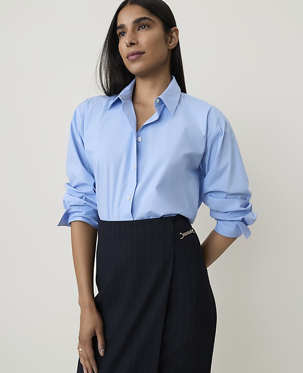 A woman in a light blue button-down shirt and a pinstripe skirt stands against a plain white background.