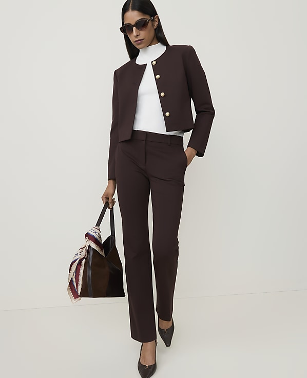 A woman in a brown suit and white turtleneck stands with a patterned scarf draped over her arm against a plain white background.