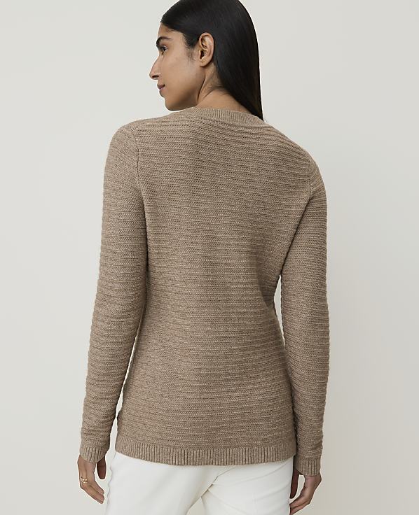 A woman in a brown textured sweater and white pants stands against a plain, light background.
